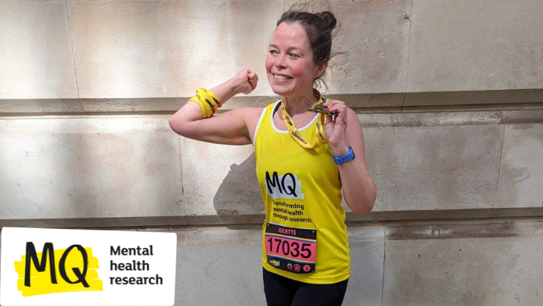 Juliette stands against a brick wall wearing a bright yellow MQ Mental Health Research running top with a race bib pinned to it. She's smiling holding a London Landmarks Half Marathon race medal.